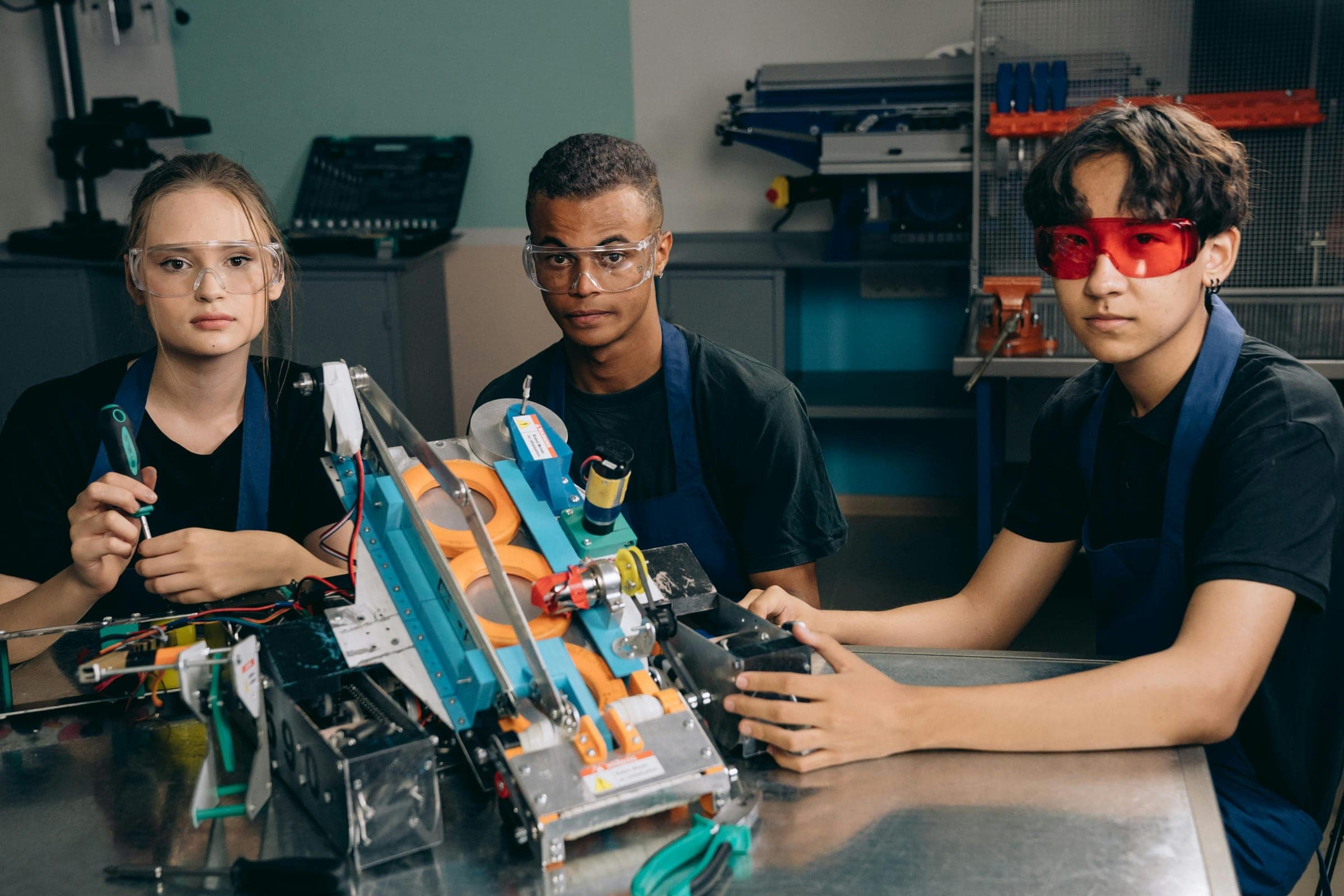 Three students in safety glasses beside a robot they have built in a workshop