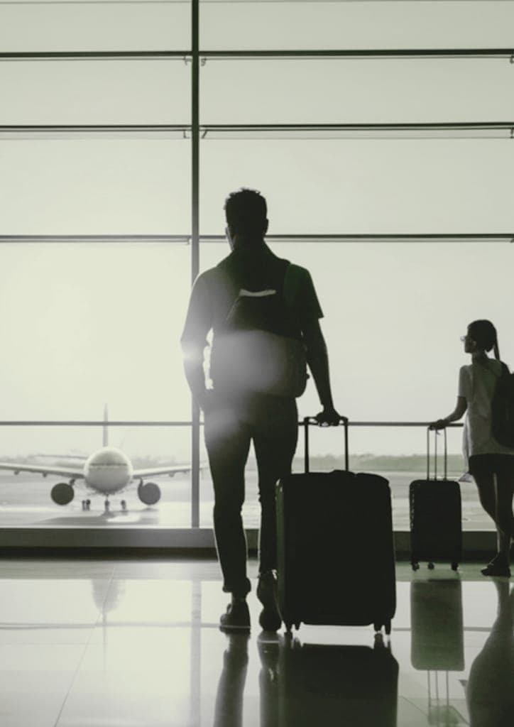 Travellers with suitcases silhouetted against an airport window, with an aircraft outside